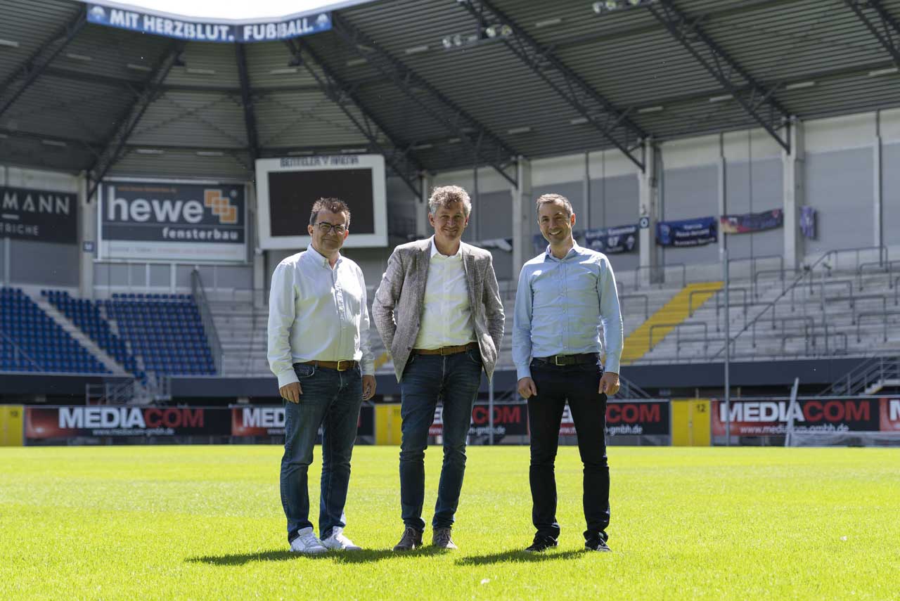 Drei Männer stehen auf dem Rasen des Stadions.