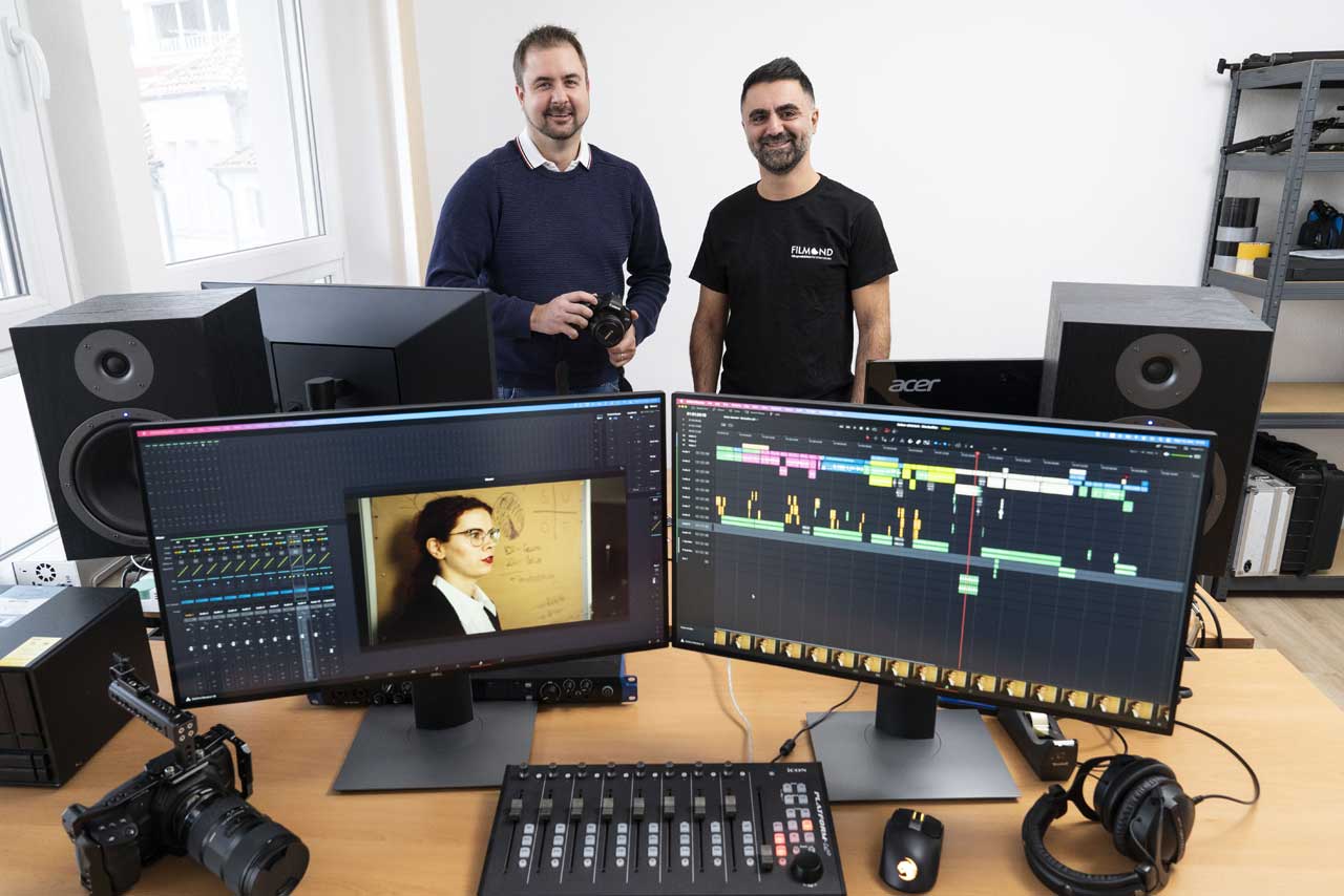 Videoschnitt, zwei Monitore und im Hintergrund zwei Männer stehend.